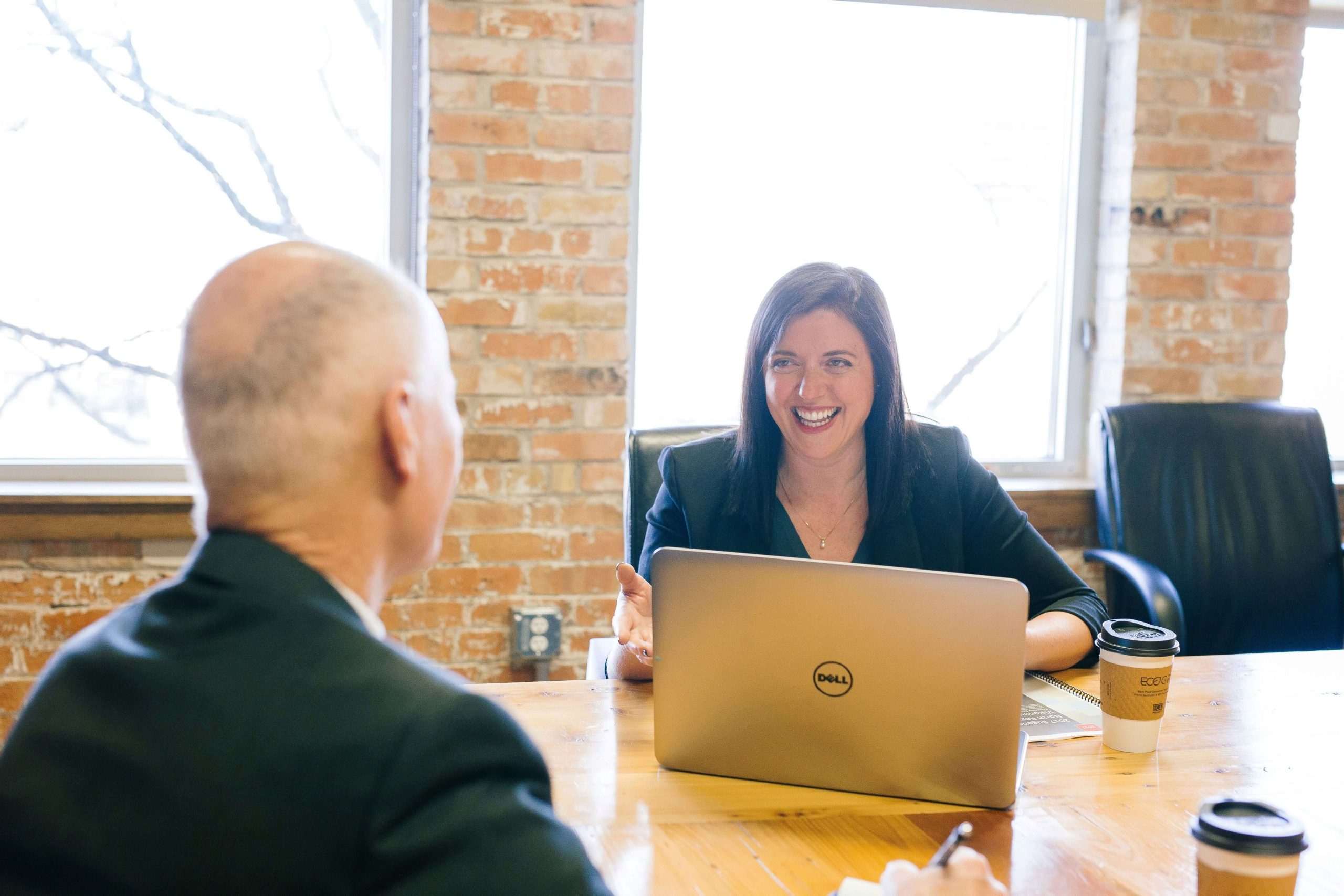 Executive Consultancy meeting between a smiling lady with a laptop and an older white male in a well-lit boardroom.