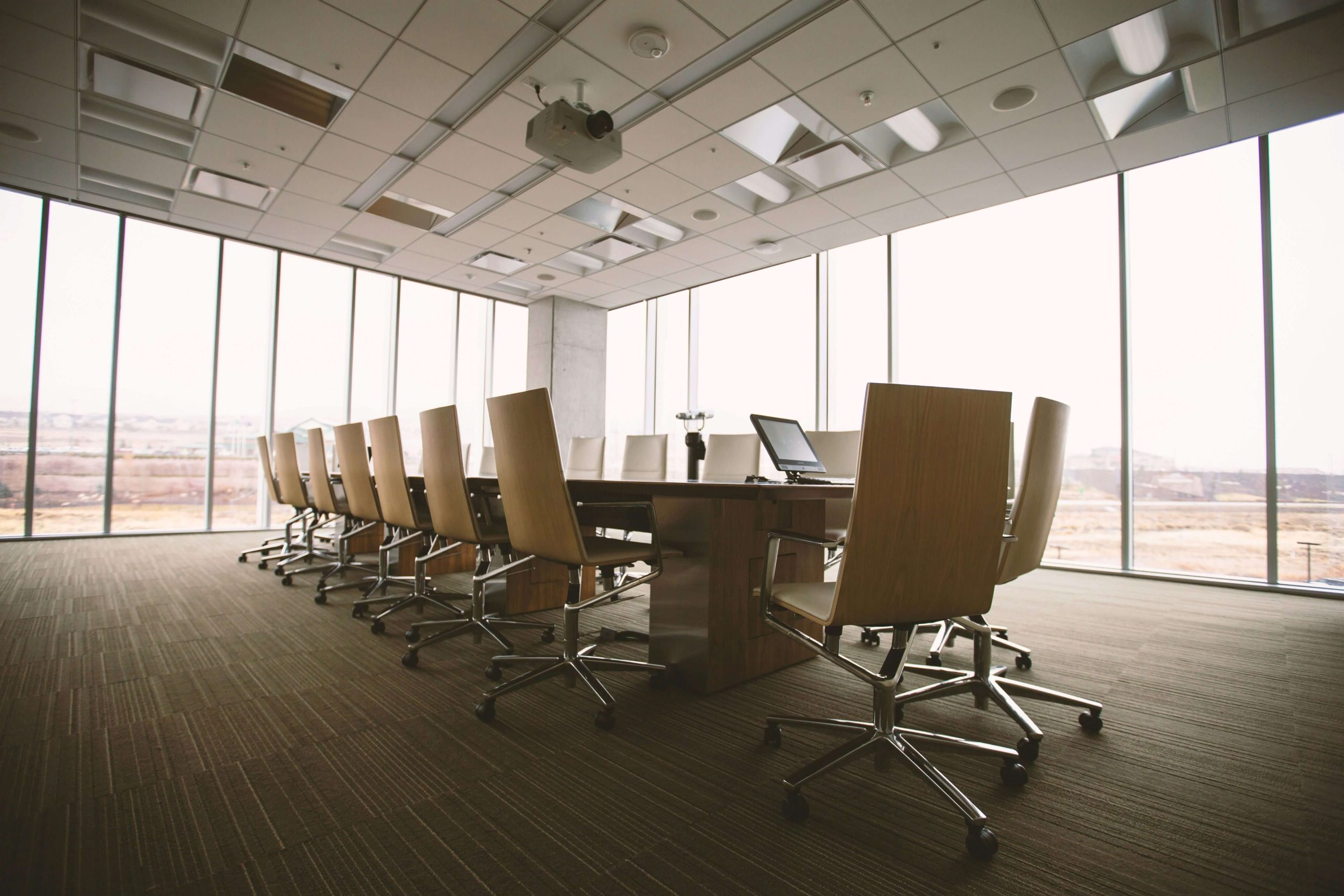 Large boardroom with transparent walls, fitted with a big rectangular table, beige chairs, and a modern overhead projector