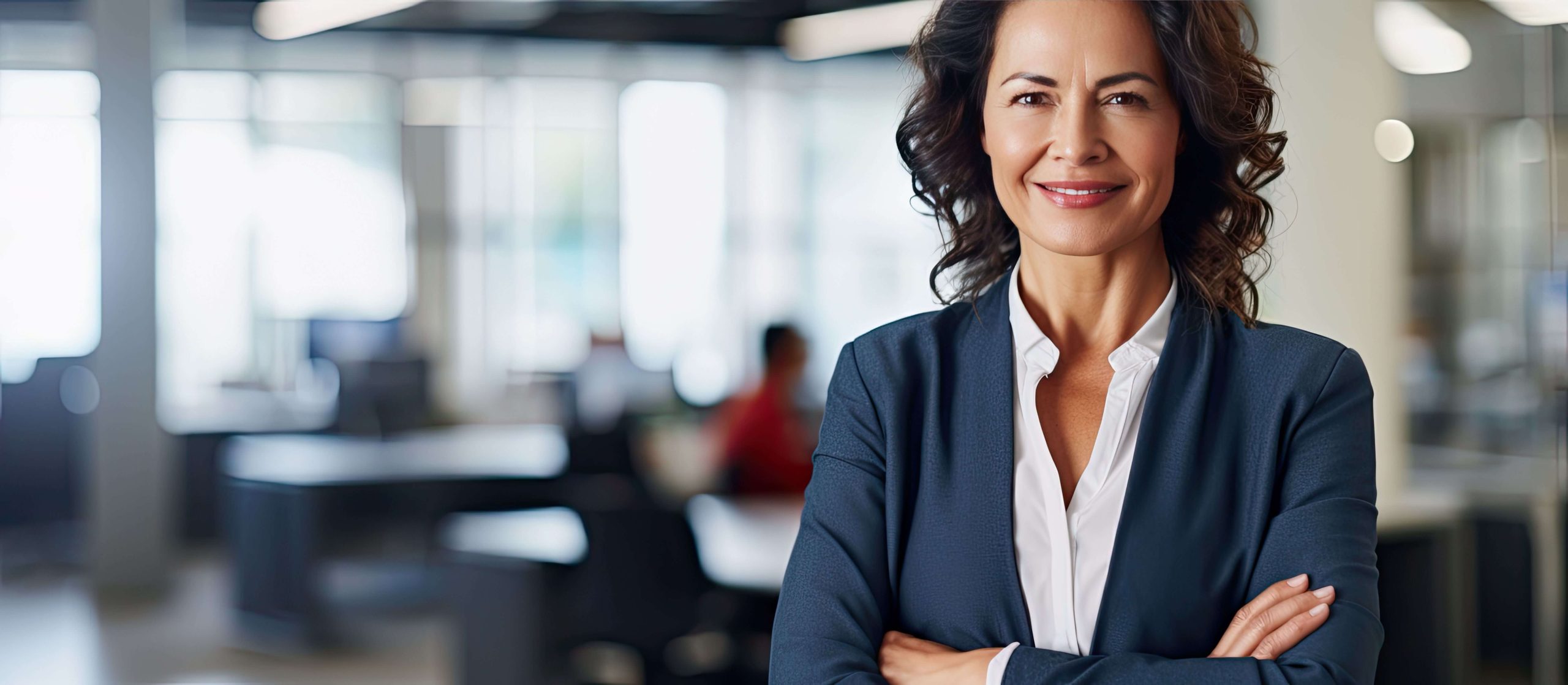 Smiling woman with curly brunette hair wearing a white shirt and a grey suit, in an office setting